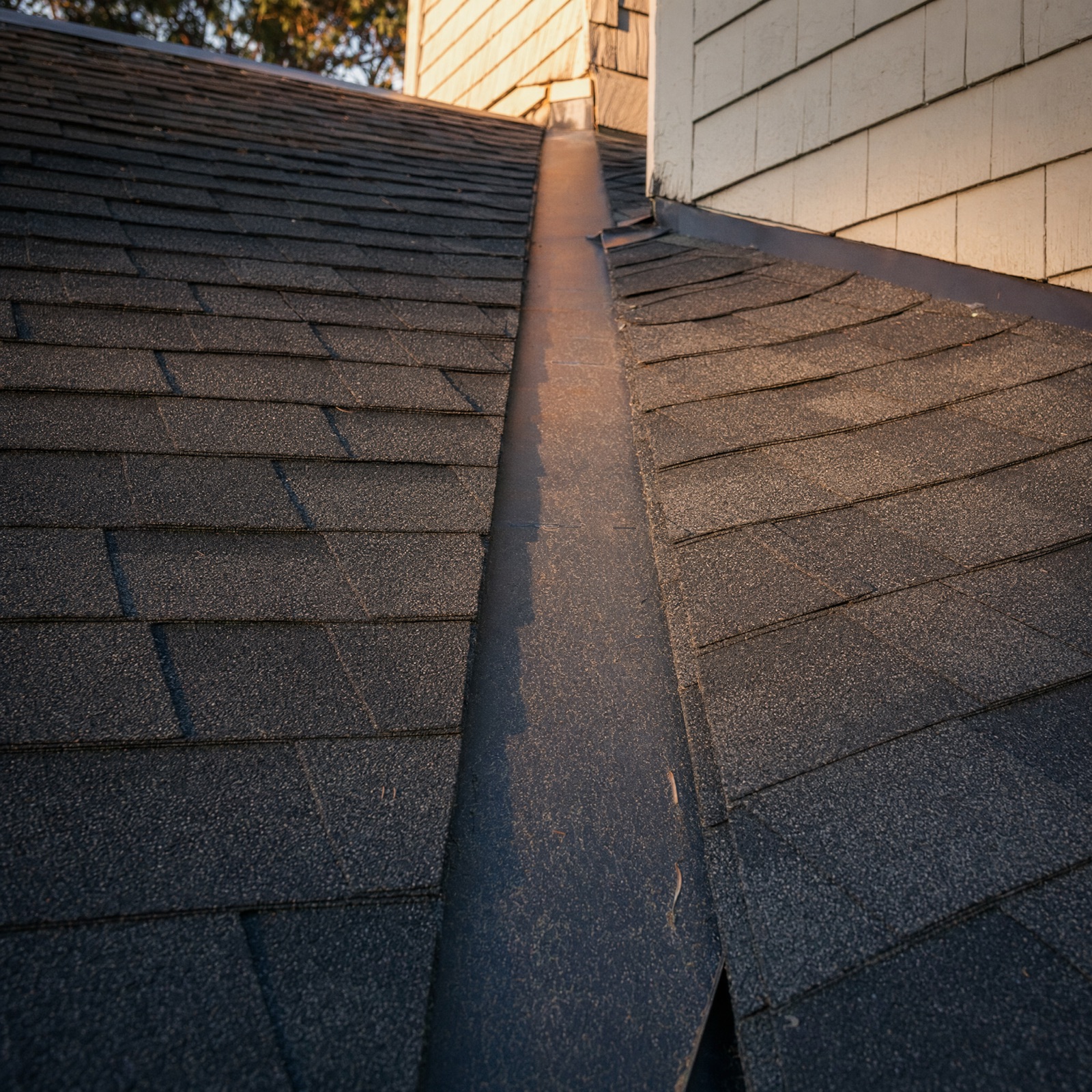 Close detail of a shingled valley in low light, the metal valley liner running straight and the shingle courses cut clean along both sides of it.