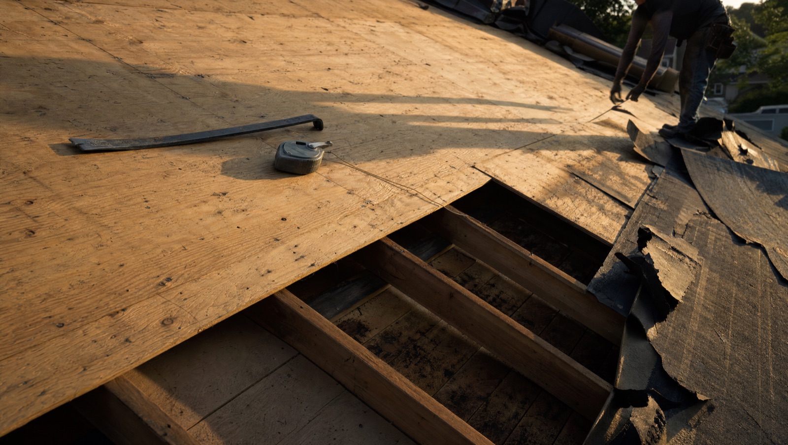 A roof stripped back to bare plywood decking with one section lifted away to show the rafters beneath, and a flat bar resting on the sound part of the slope.