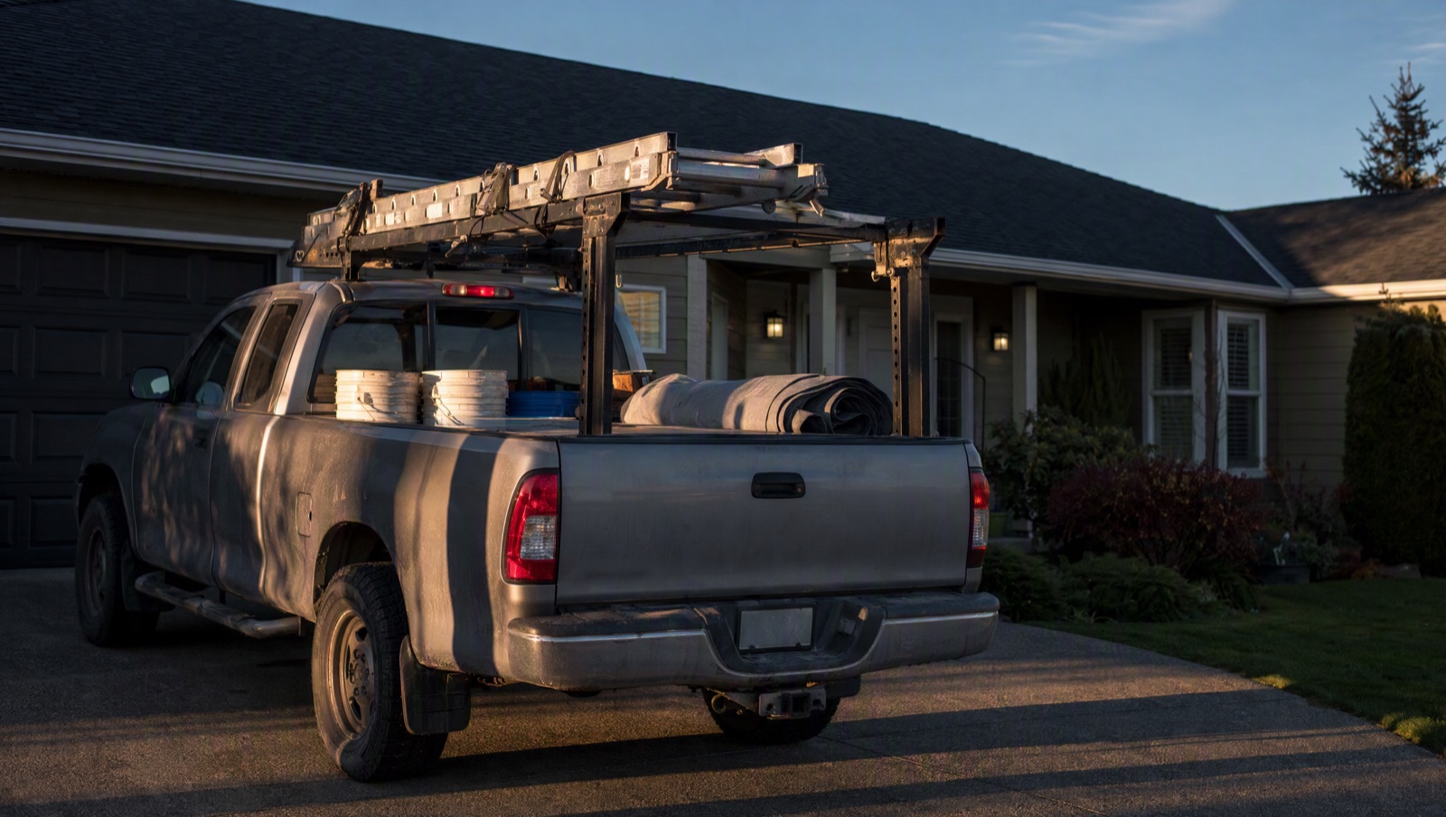 A work truck backed onto a driveway at the end of the day, ladders racked and tools stowed, a finished roofline behind it in low light.