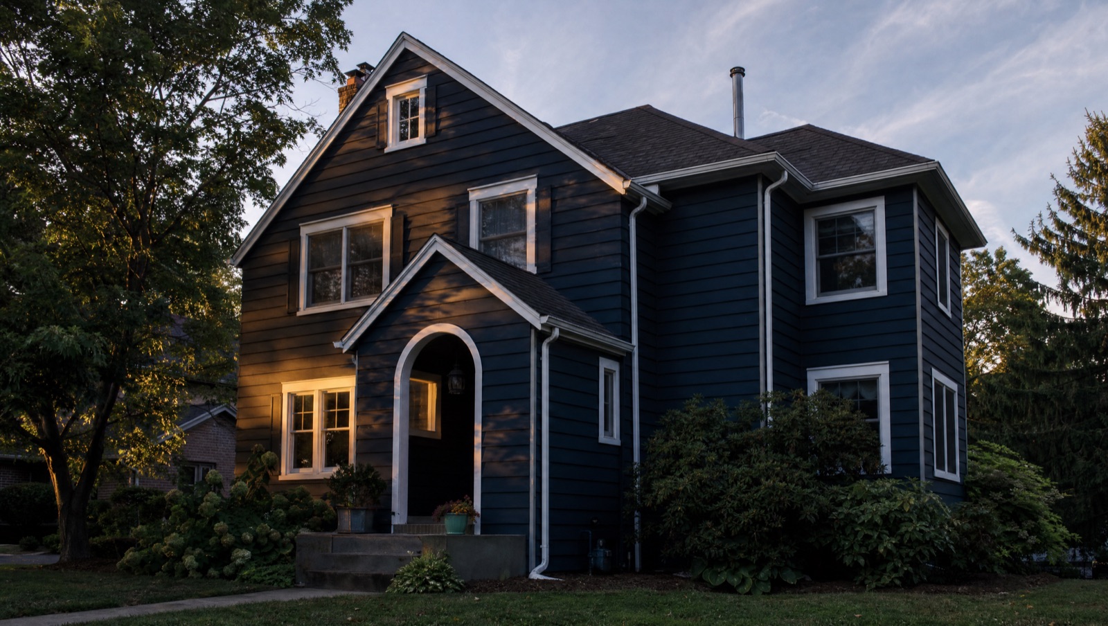 A two-storey home clad in deep navy lap siding with crisp white trim, photographed from the front lawn at dusk, warm light in the windows and mature planting at the foundation.