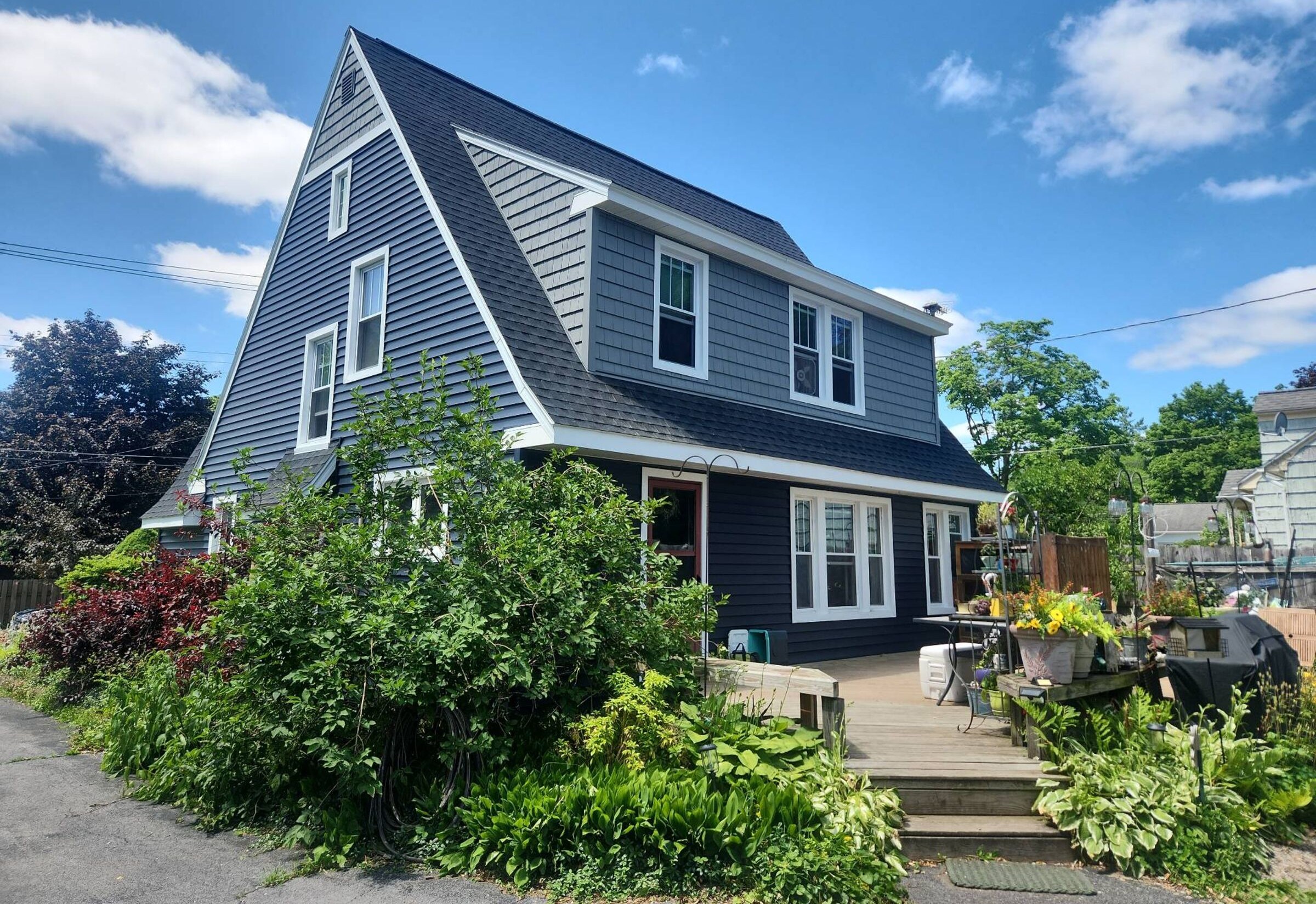 A navy-blue vinyl-sided two-storey home with a steep front gable and dormer, photographed from the front yard on a clear day with bright blue sky, multiple roof planes and a covered side porch below.