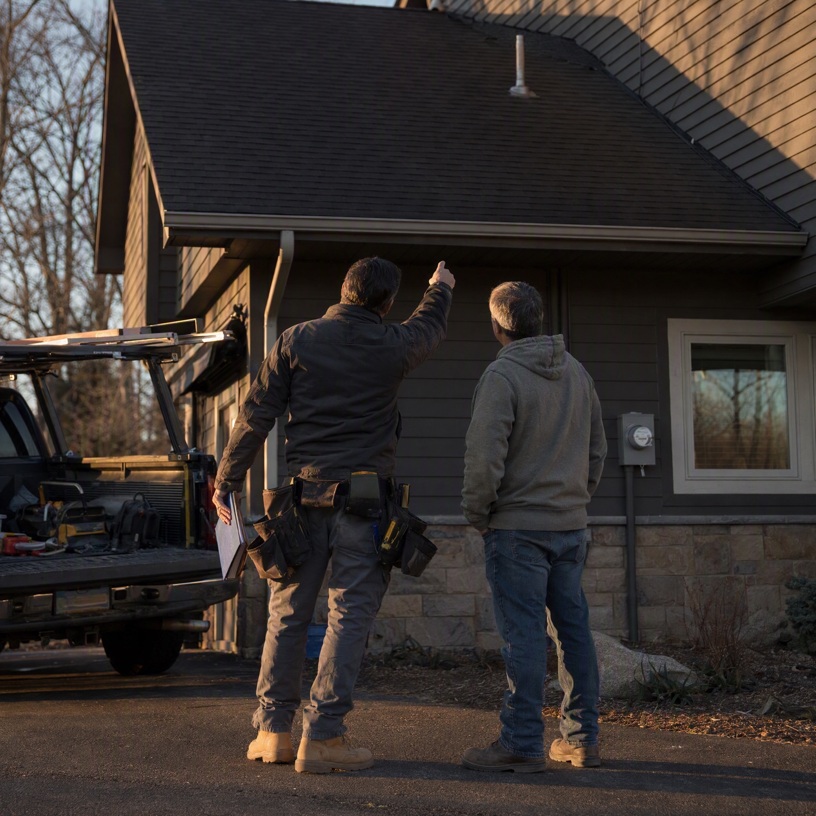A roofer in work clothes and tool belt stands with a homeowner on a driveway, gesturing up toward the eave, a work truck behind them in low evening light.