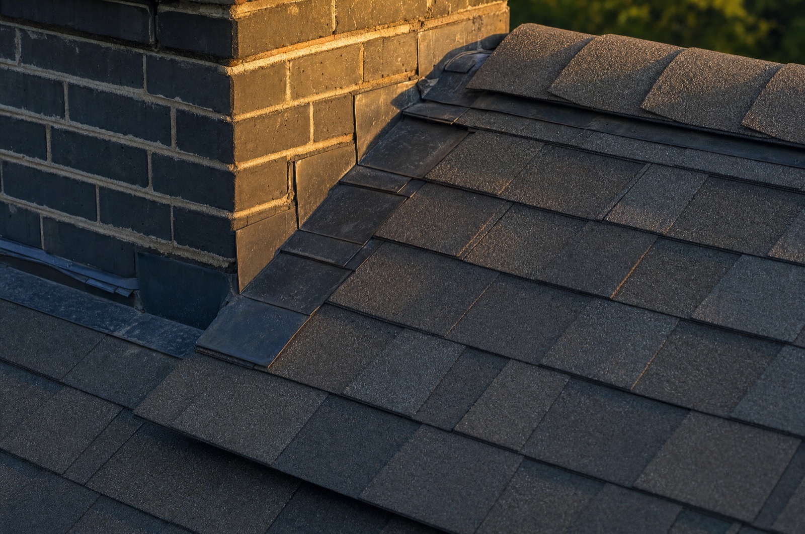 Finished roof detail where the courses meet a brick chimney: new step flashing and a counter-flashed apron tucked into the mortar joint, ridge cap running clean above.