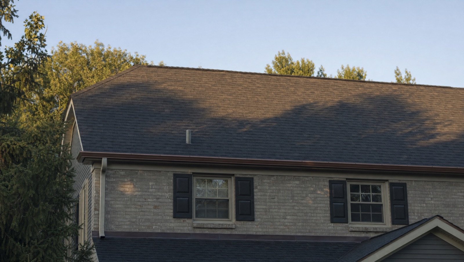 A completed asphalt shingle roof on a two-storey home at golden hour, courses running clean from eave to ridge above a brick and lap-sided elevation.