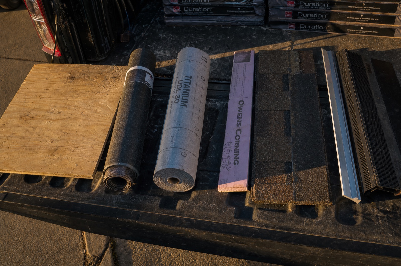 Roofing materials laid out in order on the open tailgate of a work truck: plywood decking, ice-and-water shield, synthetic underlayment, a starter strip, an architectural shingle, drip edge and a ridge vent section.