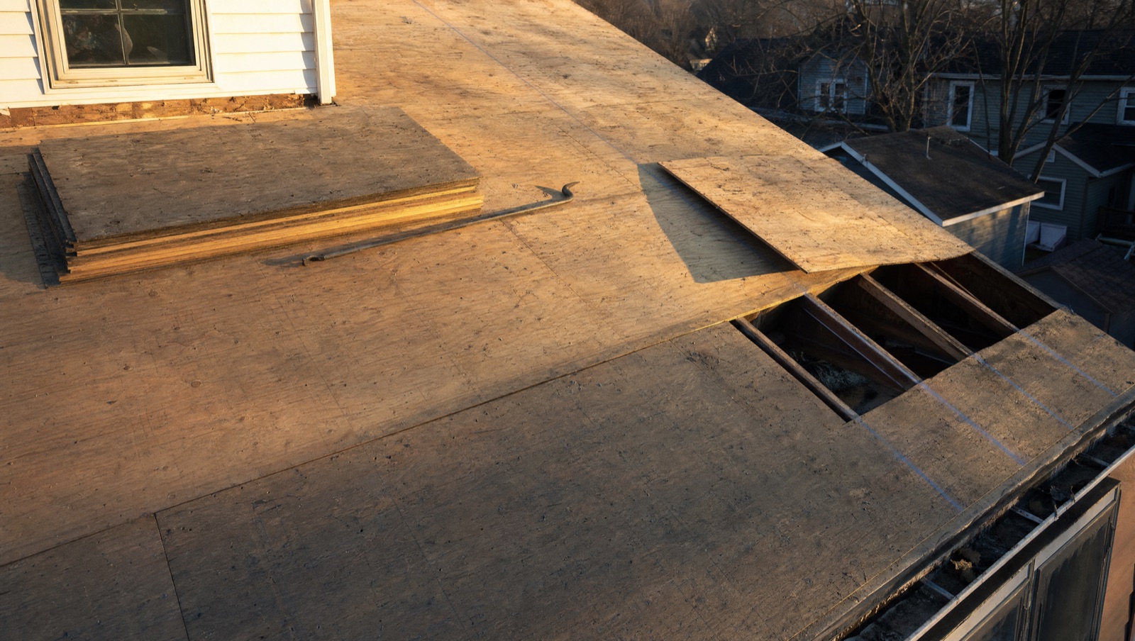 A roof stripped to bare plywood decking with one section lifted to show the rafters beneath, new decking and a flat bar resting on the sound part of the slope.