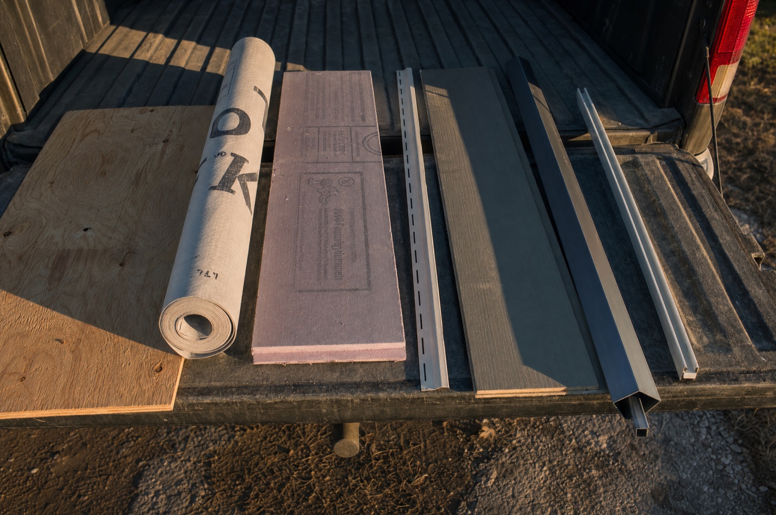 Siding materials laid out in order on the open tailgate of a work truck: sheathing, house wrap, foam board, a starter strip, lap siding, a corner post and J-channel.
