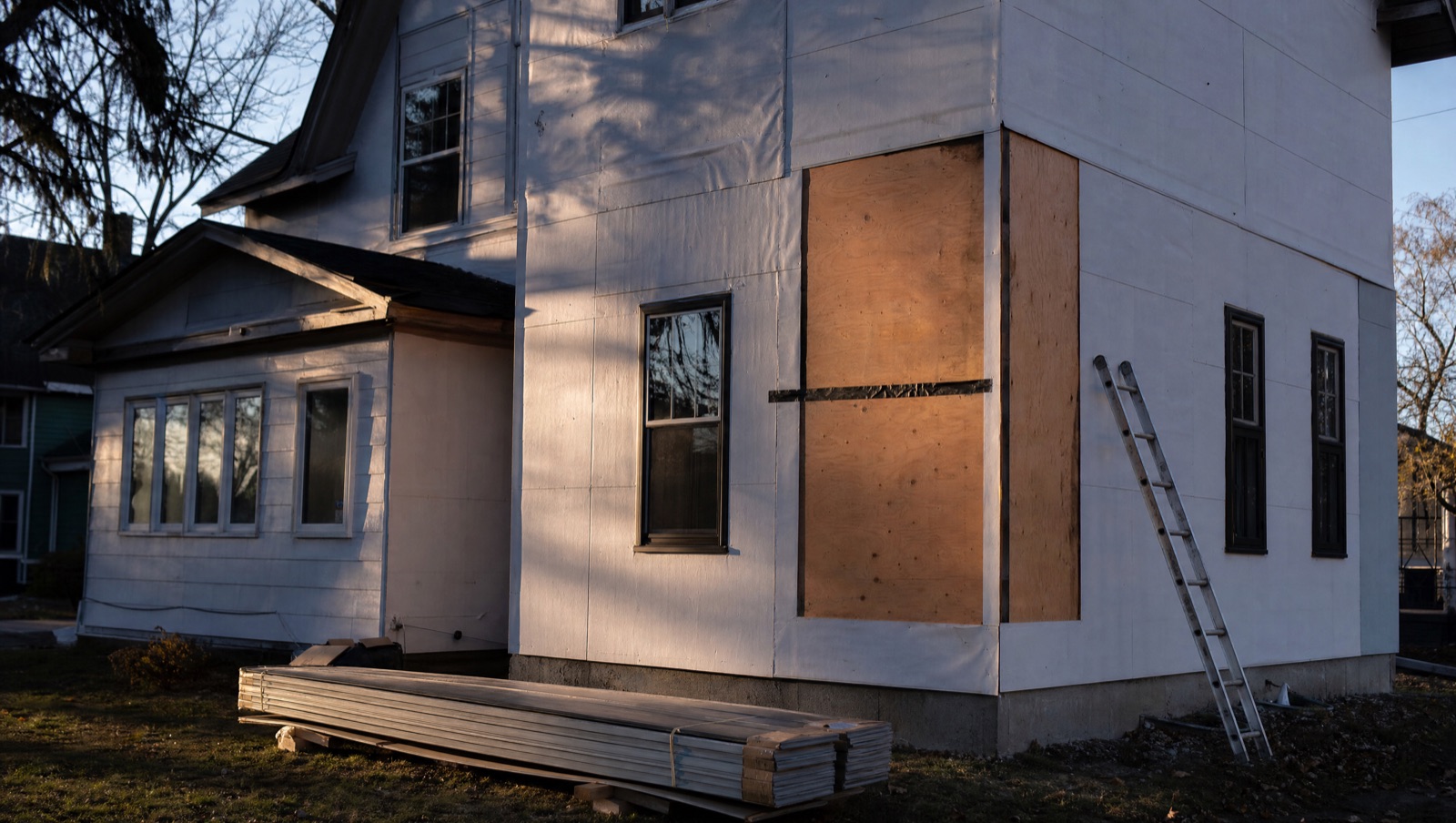 A house elevation with the old siding stripped away and plain house wrap being hung over the bare sheathing, bundles of new siding stacked on the lawn.