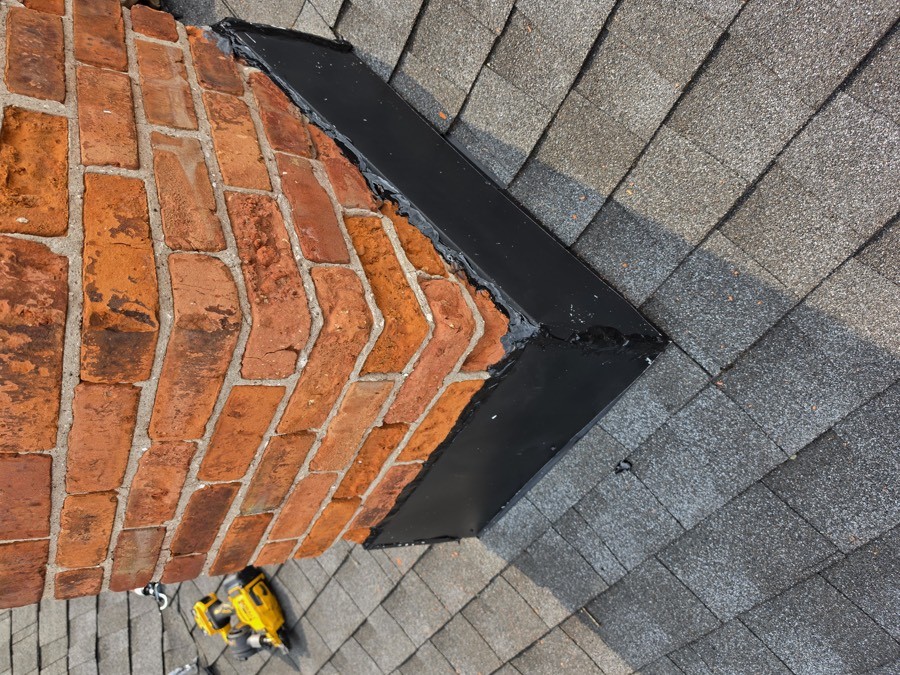 Fresh black flashing sealed around the base of a brick chimney where it meets an asphalt shingle roof, a cordless drill left on the shingles nearby.