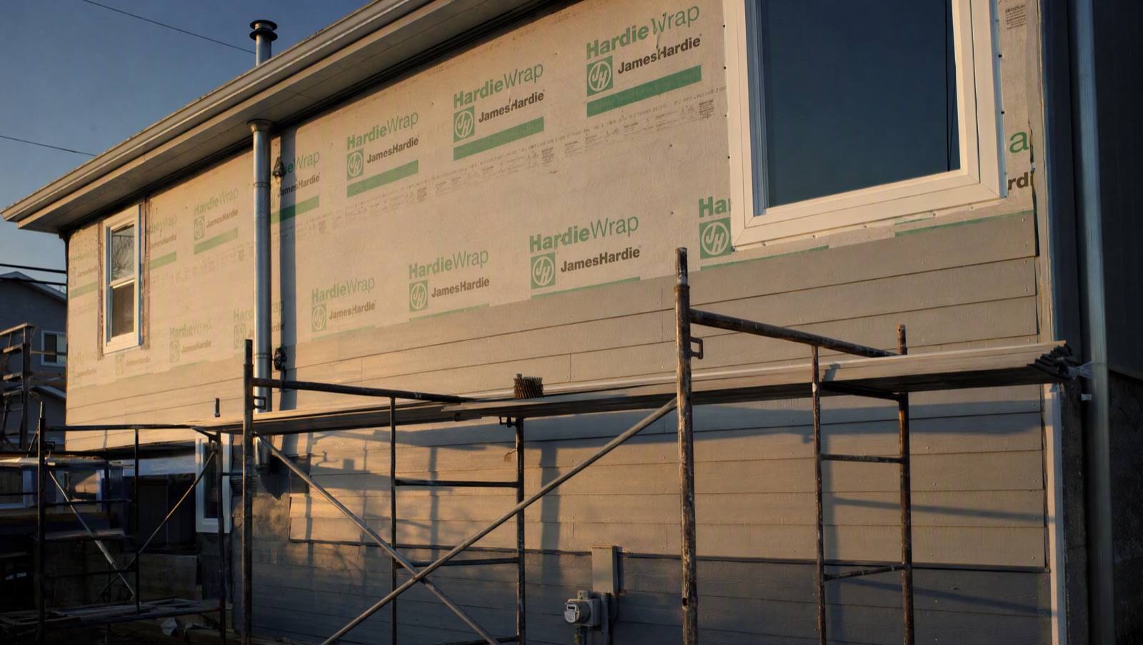 Scaffolding staged along a two-storey elevation with new grey lap siding going up course by course, gable end still open.